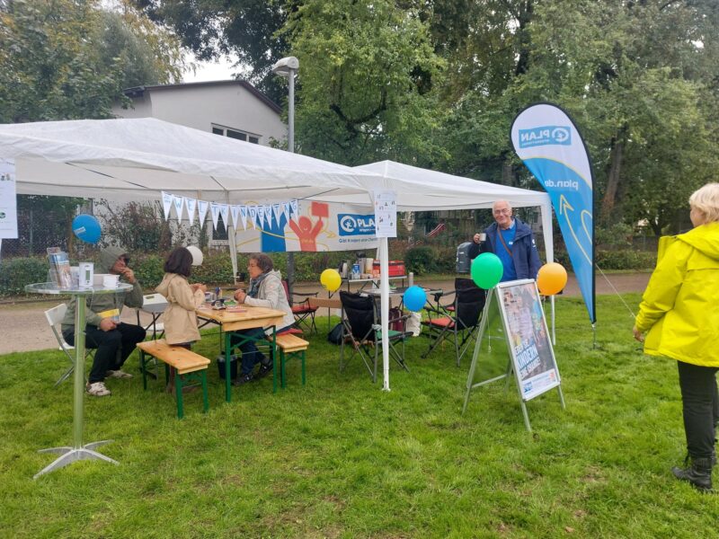 Ein Informationsstand von Plan International in einem Park, an dem Menschen an einem Tisch sitzen und ein Freiwilliger daneben steht. Auf dem Rasen stehen eine blaue Fahne und ein Schild, das für die Organisation wirbt.