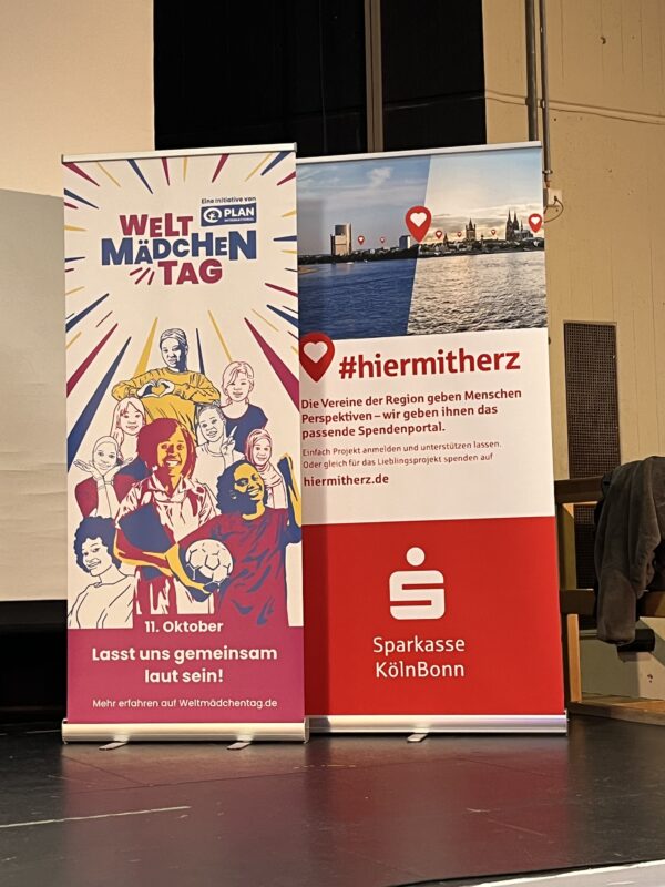 Auf einer Bühne stehen zwei vertikale Banner. Auf dem einen sind Illustrationen verschiedener Mädchen sowie der Text Welt-Mädchentag zu sehen. Das andere zeigt eine Stadtsilhouette über einem roten Sparkasse KölnBonn-Logo und dem Hashtag #hiermitherz.