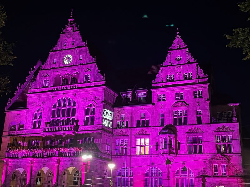 Ein kunstvoll verziertes, mehrstöckiges Steingebäude erstrahlt nachts in leuchtendem Rosa. Auf einem Banner an der Fassade stehen die Aufschriften PLAN und GIRLS GET EQUAL, während oben am linken Giebel eine Uhr zu sehen ist.