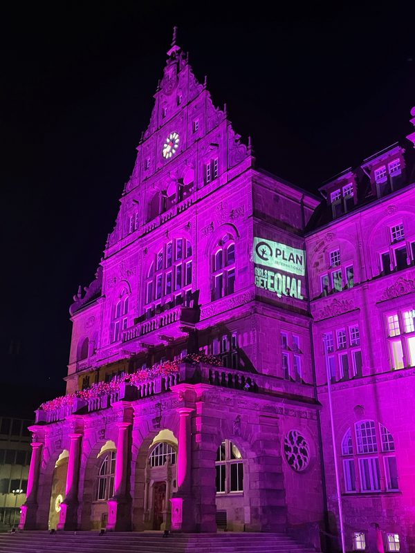 Ein historisches Steingebäude wird nachts in leuchtendem Rosa beleuchtet. Auf der Seite der Fassade ist eine große, leuchtende Projektion mit der Aufschrift Plan International, Girls Get Equal zu sehen, über einem Balkon, der mit beleuchteten Blumengirlanden geschmückt ist.