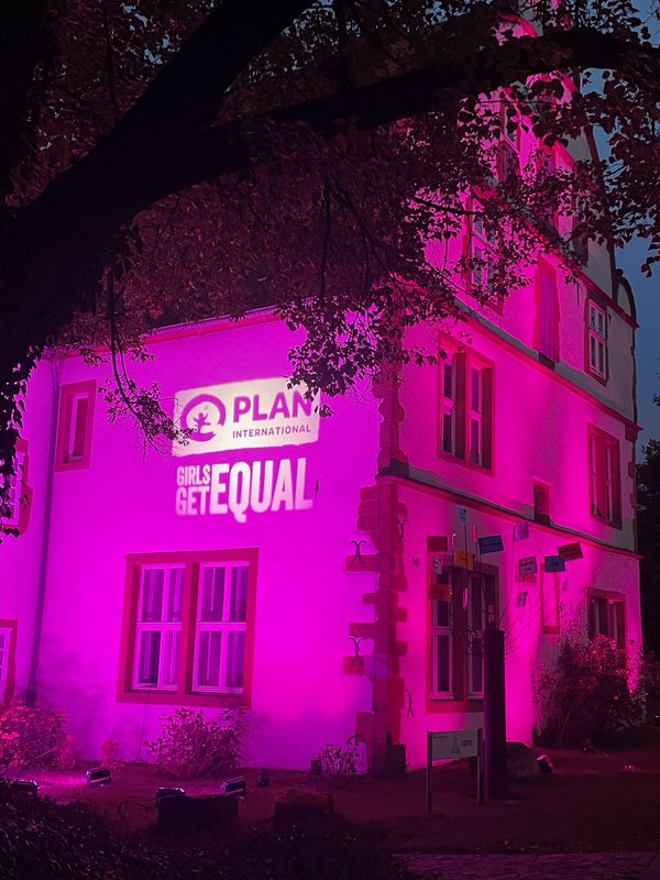 Ein Gebäude wird nachts in leuchtend rosa Licht getaucht; darauf sind das Logo von Plan International und der Schriftzug GIRLS GET EQUAL zu sehen. Bäume rahmen das Steingebäude teilweise ein, und im Vordergrund hängen kleine bunte Schilder an einem Ast.