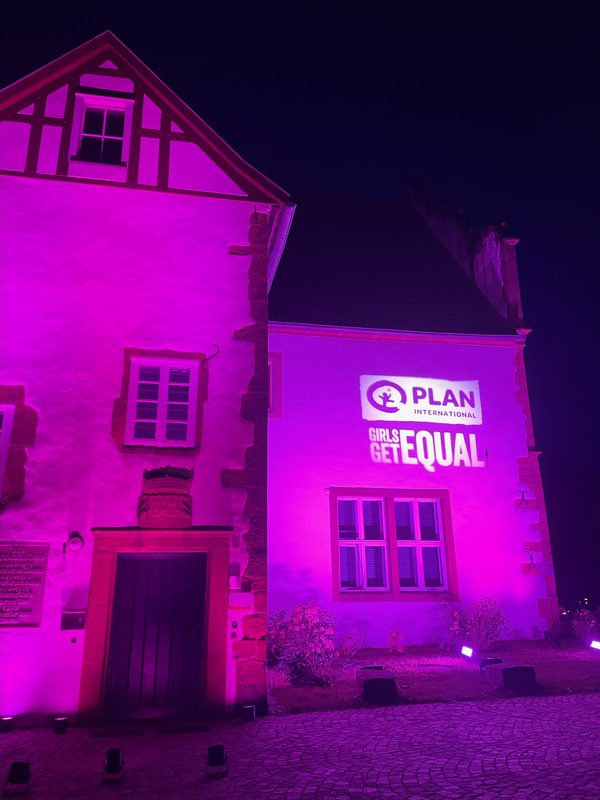 Ein historisches Steingebäude wird nachts in leuchtend rosa Licht getaucht. Eine Projektion an der Wand zeigt das Logo von Plan International über dem Schriftzug GIRLS GET EQUAL in großen, weißen Buchstaben.