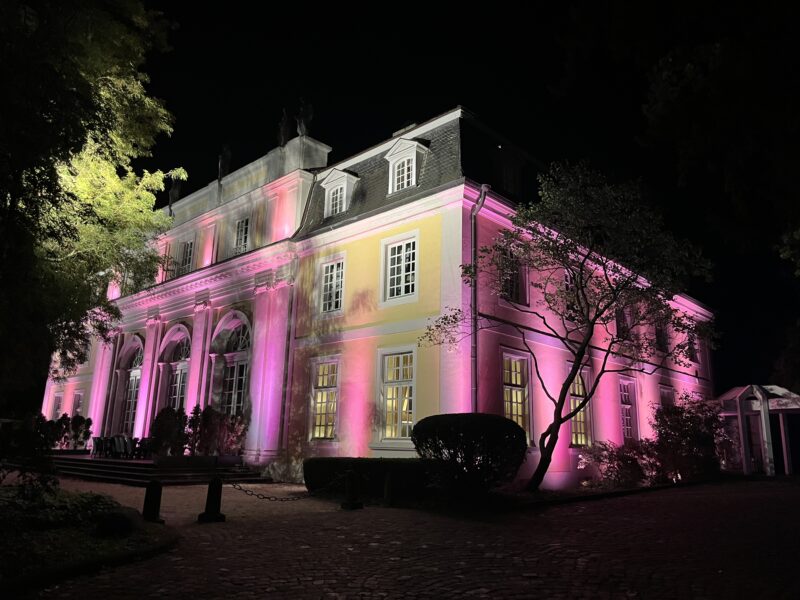 Ein historisches Herrenhaus wird nachts von leuchtend rosa und gelben Scheinwerfern beleuchtet. Das mehrstöckige Gebäude mit seinen weiß gerahmten Fenstern und dem dunklen Dach hebt sich vor einem pechschwarzen Himmel ab, während Bäume die Szene einrahmen.