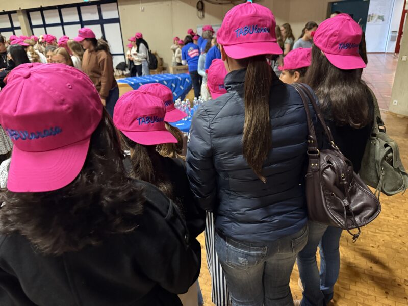 Eine Gruppe von Menschen, die von hinten zu sehen sind, trägt einheitliche, leuchtend pinkfarbene Baseballkappen, auf denen in blauer Stickerei das Wort TABUlarasa zu lesen ist. Sie stehen in einer großen Halle mit Holzboden und blicken auf einen mit einem blauen Tuch bedeckten Tisch.