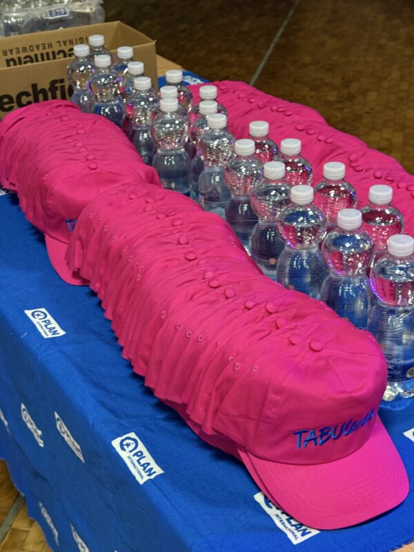 Eine lange Reihe leuchtend rosa Baseballkappen ist ordentlich auf einer blauen Tischdecke mit dem Logo von Plan International aufgestapelt. Hinter den Kappen stehen Reihen durchsichtiger Plastikwasserflaschen, und in der oberen Ecke ist ein Karton zu sehen.