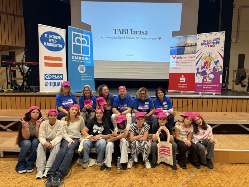 Eine Gruppe von vierzehn Frauen und Mädchen, die alle die gleichen rosa Mützen tragen, posiert auf einer Bühne. Hinter ihnen sind Banner und eine Projektionswand mit der Aufschrift TABUlarasa zu sehen. Sie lächeln vor einem Holzboden und einer Bühnenkulisse.