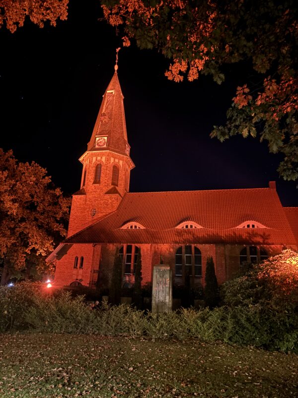 Eine Backsteinkirche mit einem hohen Kirchturm wird nachts von dramatischen roten Scheinwerfern angestrahlt. Das Gebäude ist umgeben von dunklen, sich als Silhouetten abzeichnenden Bäumen und einem Steinmonument, alles vor dem Hintergrund eines klaren, pechschwarzen Himmels.