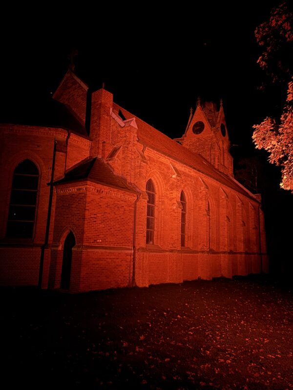 Eine Backsteinkirche wird nachts in ein grelles, leuchtend rotes Licht getaucht. Die dunklen, spitzen Fenster und der hohe Kirchturm heben sich deutlich vom pechschwarzen Himmel ab, während im Vordergrund abgefallene Blätter den Boden bedecken.