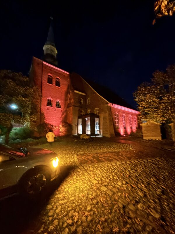 Eine nachts rot beleuchtete Backsteinkirche mit einem gewölbten Eingangsbereich und einem hohen Kirchturm. Im Vordergrund beleuchten die gelben Scheinwerfer eines Autos einen Kopfsteinpflasterweg, während eine Person in einer gelben Jacke in der Nähe des Eingangs zum Gebäude steht.