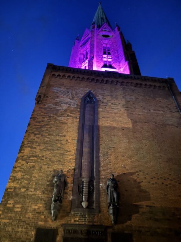 Ein hoher Kirchturm aus Backstein wird von unten durch ein leuchtendes Magenta-Licht vor dem Hintergrund eines tiefblauen Nachthimmels beleuchtet. Zwei dunkle Statuen stehen zu beiden Seiten eines vertikalen architektonischen Akzents an der unteren Steinfassade.