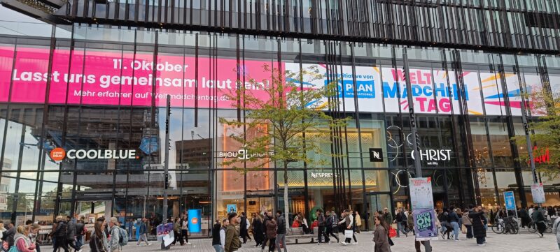 Ein modernes Einkaufszentrum mit Glasfassade zeigt große Banner, die für den Internationalen Mädchentag werben. Die Passanten spazieren auf dem Bürgersteig vor dem Gebäude entlang, an dem sich Ladenfronten von Marken wie Coolblue, Nespresso und Christ befinden.