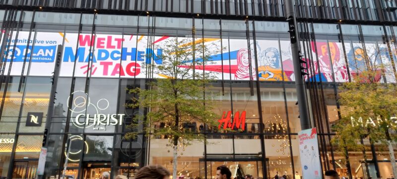 An der Glasfassade eines modernen Gebäudes ist ein großes Banner zum Welt-Mädchentag mit farbenfrohen Illustrationen verschiedener Mädchen zu sehen. Darunter sind hinter Bäumen an einer Stadtstraße die Schaufenster von Christ, H&M und Mango zu erkennen.