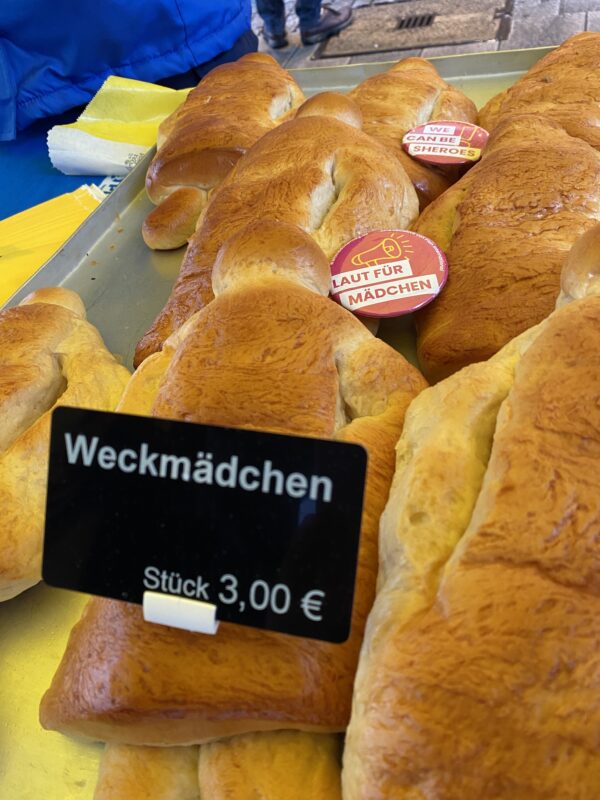 Auf einem Metalltablett liegen mehrere goldbraune, puppenförmige süße Brötchen. Ein schwarzes Preisschild weist sie als Weckmädchen für 3,00 € aus, während auf einigen Brötchen kleine runde Aufkleber mit den Aufschriften We can be sheroes und Laut für Mädchen zu sehen sind.