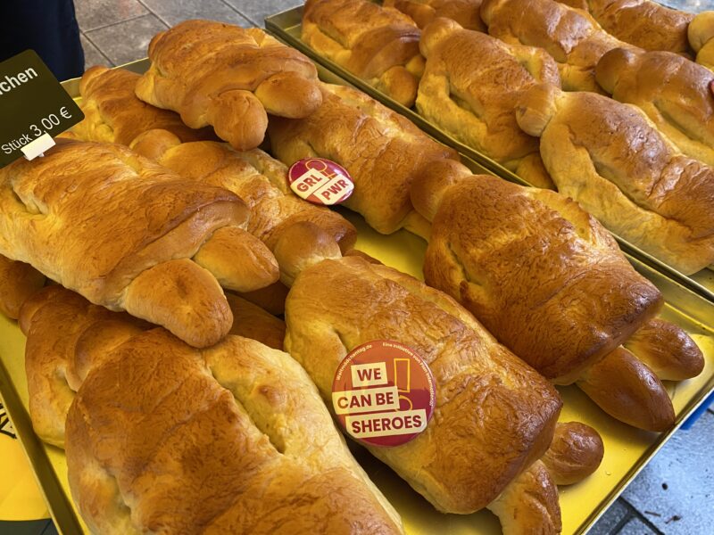 Auf Metalltabletts liegen goldbraune, schildkrötenförmige Brotlaibe. Zwei der Laibe sind mit runden Aufklebern versehen, auf denen GRL PWR und WE CAN BE SHEROES steht. Ein kleines Schild in der Ecke gibt den Preis von 3,00 Euro pro Stück an.