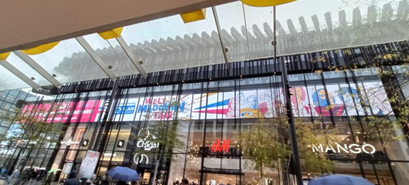 Die Fassade eines modernen Einkaufszentrums ist mit großen Glasfenstern versehen, an denen bunte Werbebanner zum Welt-Mädchentag hängen. Unterhalb der Banner sind die beleuchteten Ladenschilder von H&M und Mango zu sehen, während an einem regnerischen Tag Menschen mit Regenschirmen daran vorbeigehen.