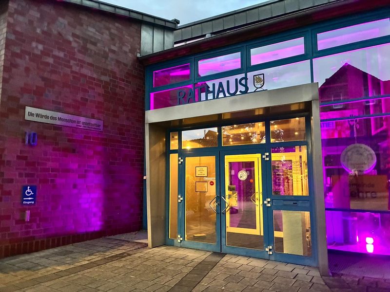 Der Eingang zu einem Backsteingebäude mit der Aufschrift RATHAUS ist in helles rosa Licht getaucht. An der Wand links ist ein Schild mit der Aufschrift Rollstuhlzugang angebracht, neben einer Gedenktafel mit dem deutschen Text: Die Würde des Menschen ist unantastbar.