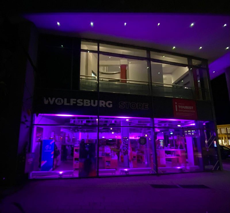 Der Wolfsburg Store bei Nacht, beleuchtet von leuchtend violetten Innenleuchten. Das Gebäude zeichnet sich durch große Glasfenster, eine dunkle Fassade mit auffälliger weißer Beschriftung und ein rotes Schild der Touristeninformation in der Ecke aus.