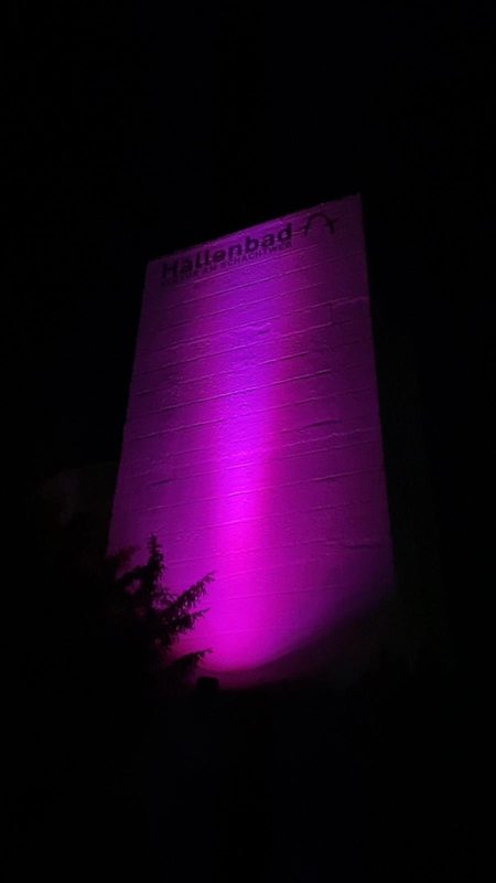 Ein hoher Turm, der nachts von einem hellen, vertikalen Streifen in Magenta beleuchtet wird. Oben am Turm ist der Schriftzug Hallenbad Kultur am Schachtweg in Schwarz zu sehen, unten links sind die dunklen Silhouetten von Ästen zu erkennen.