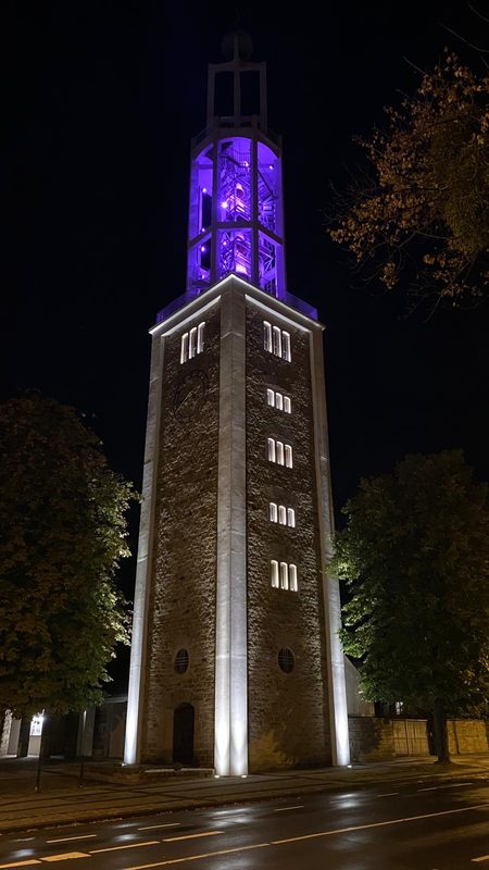 In der Nacht ragt ein hoher Uhrturm aus Stein empor, der von unten durch helles weißes Licht und von oben durch violettes Licht aus dem Glockenturm beleuchtet wird. Der Turm wird von Bäumen flankiert, und im Vordergrund verläuft eine dunkle Straße.