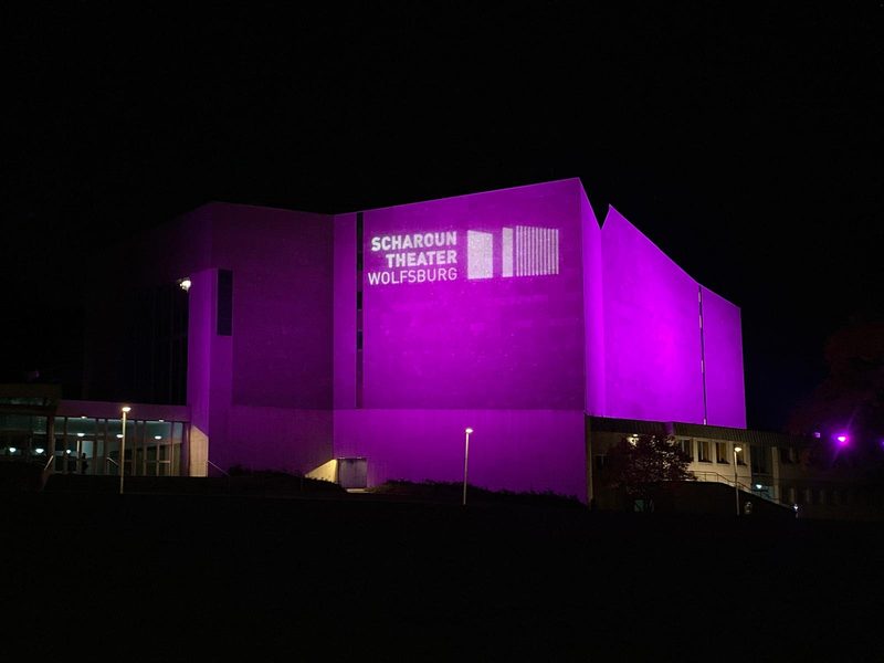 Das Scharoun-Theater Wolfsburg bei Nacht, beleuchtet von einer leuchtend violetten Projektion. Der Name des Gebäudes und ein grafisches Motiv werden in weißem Licht an die Wand projiziert und heben sich deutlich von der dunklen, umgebenden Landschaft ab.