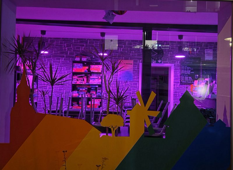 Der Innenraum eines Ladengeschäfts wird von violetten Neonröhren beleuchtet, die die Regale mit Brettspielen vor einer dunklen Steinwand in Szene setzen. Im Vordergrund sind bunte Papiersilhouetten einer Windmühle, von Bäumen und Gebäuden auf die Scheibe geklebt.