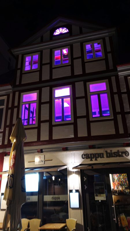 Ein dreistöckiges Fachwerkhaus bei Nacht, aus dessen oberen Fenstern leuchtendes Magenta strahlt. Darunter ist die Fassade des Cappu Bistro zu sehen, mit einem zusammengeklappten Sonnenschirm und gelben Stühlen auf dem Bürgersteig.