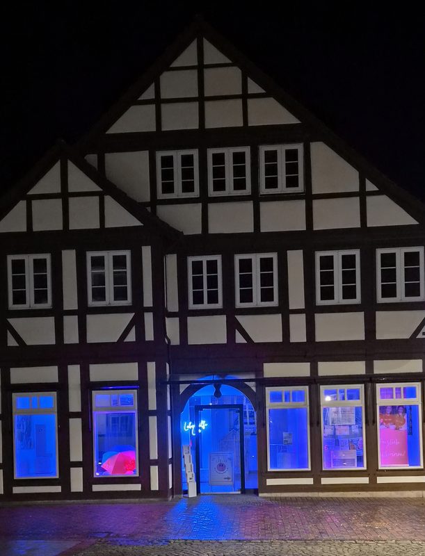 Ein Fachwerkhaus wird nachts von hellblauem Licht aus den Fenstern im Erdgeschoss beleuchtet. Das Gebäude hat einen dunklen Holzrahmen vor weißen Wänden, und auf einem kleinen rosa Schild im vorderen Fenster steht Herzlich Willkommen.