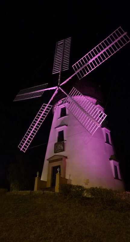 Eine historische weiße Windmühle ragt vor einem pechschwarzen Nachthimmel empor und wird von leuchtend rosa Scheinwerfern hell beleuchtet. Ihre großen, gitterartigen Flügel sind diagonal geneigt und tauchen das Bauwerk und das dunkle Gras darunter in ein lebhaftes Licht.