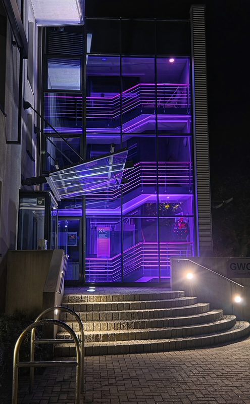 Der Eingang eines modernen Gebäudes bei Nacht besticht durch eine glasverkleidete Treppe, die von leuchtend violetten Neonlichtern beleuchtet wird. Steinstufen führen hinauf zum Eingangsbereich, wo durch das Glas ein leuchtendes Schild und eine Person in einer roten Jacke zu sehen sind.