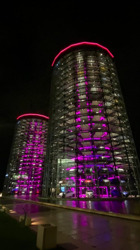 Zwei gläserne, zylinderförmige Parkhäuser, die von leuchtend violetten LED-Lichtern beleuchtet werden, ragen vor dem dunklen Nachthimmel empor. In den transparenten Bauwerken sind Reihen von Autos zu sehen, die ihr magentafarbenes Leuchten auf den nassen Asphalt darunter werfen.