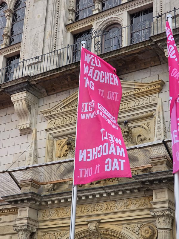 Vor einem historischen Steingebäude mit Bogenfenstern und geschnitzten Löwenreliefs hängen leuchtend rosa vertikale Banner. Auf den Fahnen ist in weißer Schrift auf Deutsch für den Welt-Mädchentag bzw. den Internationalen Tag des Mädchens am 11. Oktober geworben.