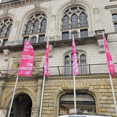 Vor einem historischen, mehrstöckigen Steingebäude mit kunstvoll verzierten Bogenfenstern und einer mit Schnitzereien verzierten Fassade stehen vier leuchtend rosa Fahnen mit weißer Aufschrift zum Internationalen Mädchentag.
