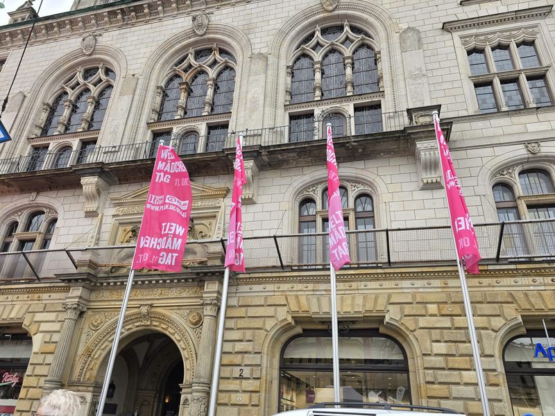 Vor einem historischen, mehrstöckigen Steingebäude mit kunstvoll verzierten Bogenfenstern und einer mit Schnitzereien verzierten Fassade stehen vier leuchtend rosa Fahnen mit weißer Aufschrift zum Internationalen Mädchentag.