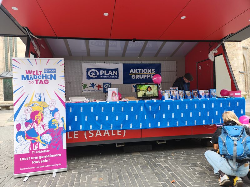 Auf einem mit Kopfsteinpflaster gepflasterten Platz steht ein Informationsstand von Plan International, an dem ein großes, hochformatiges Banner zum Welt-Mädchentag zu sehen ist. Hinter einem blauen Tisch, der mit Werbematerialien geschmückt ist, steht ein Mann, während in der Nähe eine Person mit einem blauen Rucksack kniet.