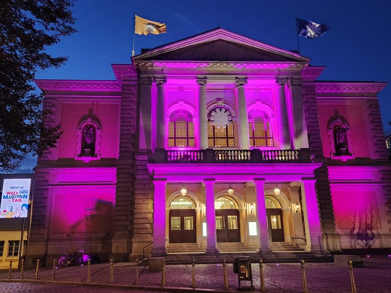 Ein prächtiges neoklassizistisches Gebäude wird in der Abenddämmerung in leuchtendes violettes Licht getaucht. Vom Dach wehen zwei Fahnen, und auf einem Schild links steht Heute ist Weltmädchentag. Auf dem gepflasterten Platz vor der Steintreppe steht ein Fahrrad.