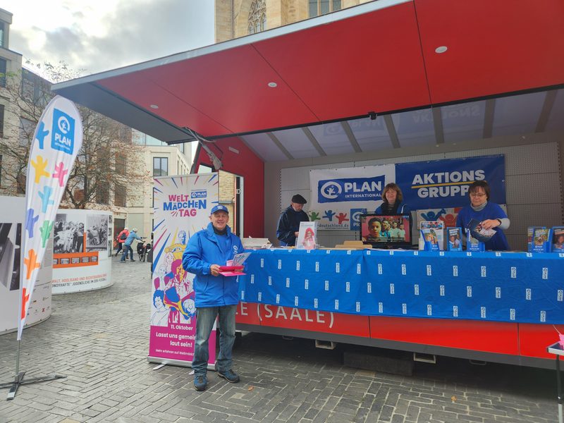 Ein Mann in blauer Jacke und Mütze steht vor einem rot-weißen Werbestand von Plan International. Hinter der Theke arbeiten zwei Frauen unter einem Banner mit dem Logo der Organisation, während ein daneben stehendes Hochformat-Schild für den Internationalen Tag des Mädchens wirbt.