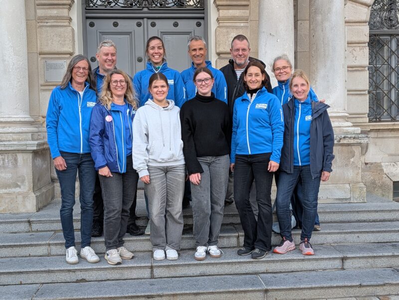 Eine Gruppe von elf Erwachsenen posiert auf einer Steintreppe vor dem Eingang eines grauen Gebäudes. Die meisten tragen einheitliche hellblaue Jacken mit PLAN-Logos, während andere legere Pullover oder Mäntel tragen; alle lächeln in die Kamera.