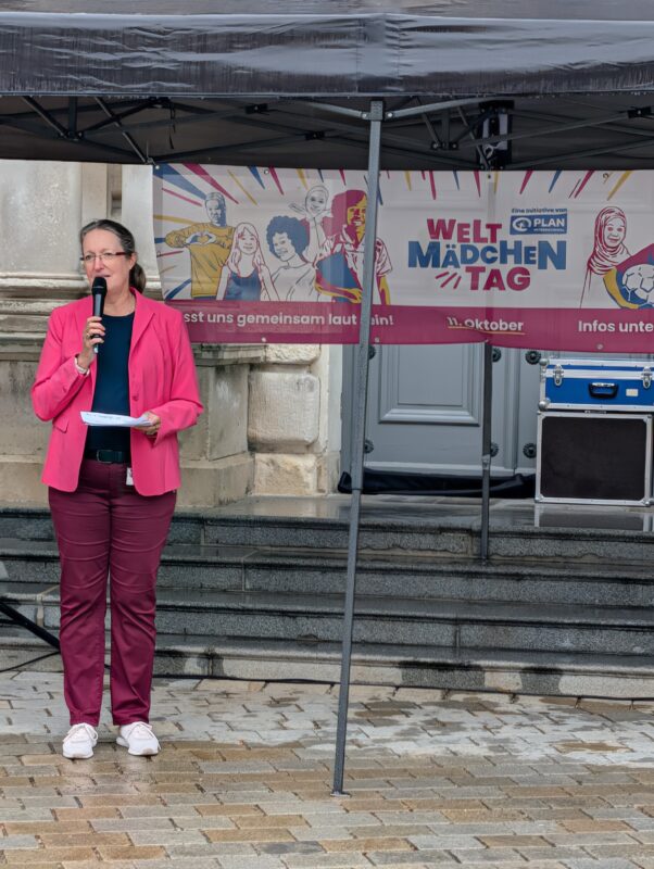 Eine Frau mit Brille, die einen rosa Blazer trägt, spricht vor einem Banner zum Internationalen Tag der Mädchen in ein Mikrofon. Sie steht auf einem steinernen Platz unter einem dunklen Vordach, hinter ihr befinden sich der Eingang eines Steingebäudes und eine Tonanlage.