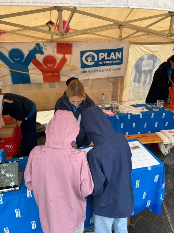 Zwei Personen in Kapuzenjacken stehen an einem Stand von Plan International. Sie stehen einem Mitarbeiter gegenüber, der hinter einem mit blauem Stoff bezogenen Tisch steht, auf dem Flyer und Broschüren ausliegen, unter einem großen Vordach mit dem Logo und dem Markenauftritt der Organisation.