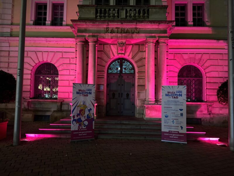 Ein historisches Rathausgebäude wird nachts in leuchtend rosa Licht getaucht, und vor der Steintreppe stehen zwei identische Werbebanner zum Internationalen Mädchentag. Die Banner sind mit farbenfrohen Illustrationen und deutschen Texten versehen.