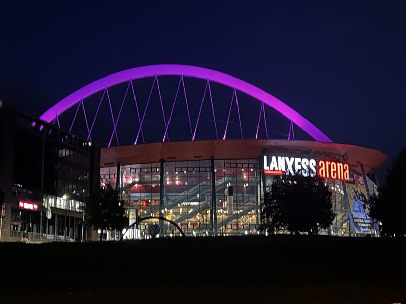 Die LANXESS Arena bei Nacht, beleuchtet von einem leuchtend violetten Bogendach. Die große Glasfassade des Stadions strahlt in warmem Licht, und das LANXESS-Arena-Logo ist in roten und weißen Buchstaben vor dem dunkelblauen Himmel zu sehen.