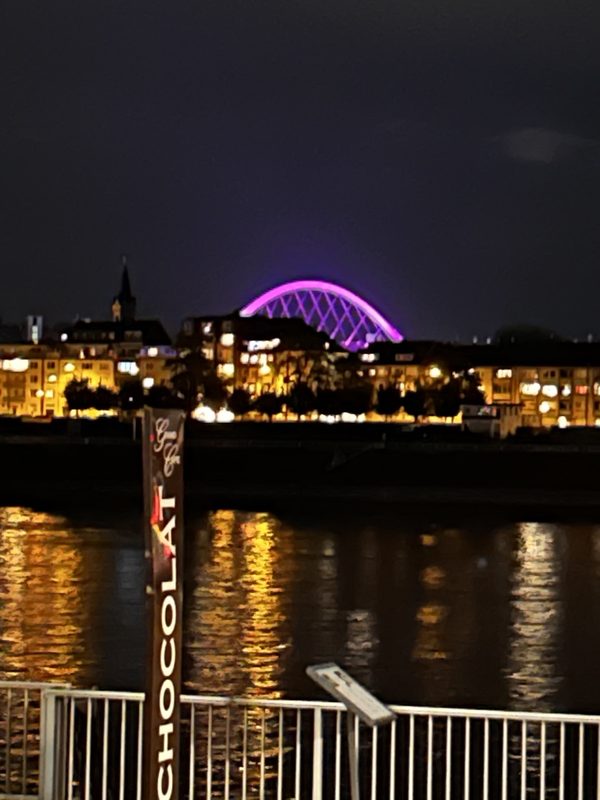 Eine leuchtend violette Bogenbrücke erstreckt sich nachts über den Horizont und hebt sich als Silhouette vom dunklen Himmel ab. Im Vordergrund steht ein Schild mit der Aufschrift CHOCOLAT in der Nähe eines Flusses, in dessen plätscherndem Wasser sich die Lichter der Stadt spiegeln.