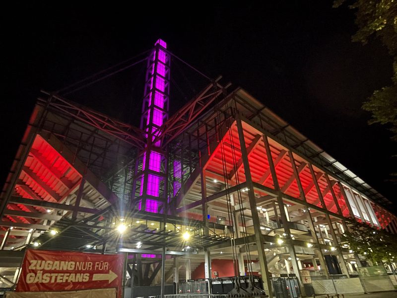 Ein großes Stadion bei Nacht: Im Mittelpunkt steht ein hellviolett beleuchteter Turm, dessen stufenförmige Tribünen in leuchtendem Rot erstrahlen. Im Vordergrund ist ein rotes Transparent mit der Aufschrift ZUGANG NUR FÜR GÄSTEFANS zu sehen, auf dem ein weißer Pfeil nach rechts zeigt.