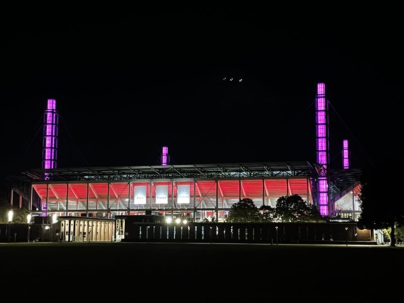 Ein großes Stadion wird nachts durch leuchtend rote Beleuchtung entlang der Dachlinie und hohe, violett leuchtende Säulen an den Ecken beleuchtet. Das Bauwerk hebt sich vom dunklen Himmel ab, während sanfte Außenbeleuchtung das umliegende Gelände in Licht taucht.