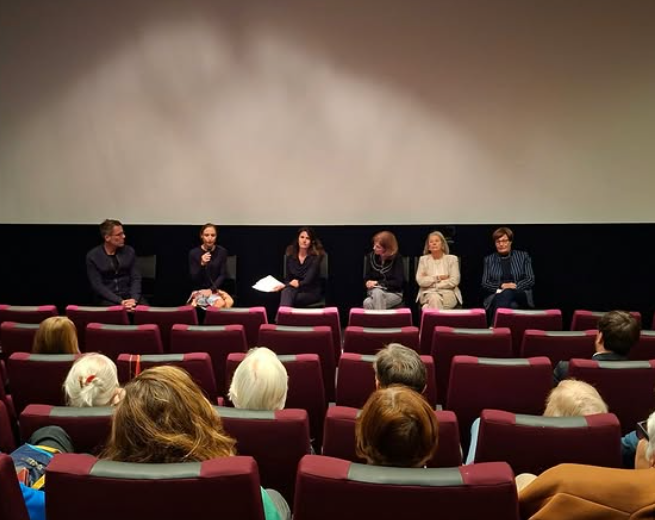 Eine sechsköpfige Diskussionsrunde sitzt auf einer Bühne vor einer leeren Leinwand und spricht zu einem Publikum, das auf dunkelroten Theatersesseln sitzt. Die Diskussionsteilnehmer tragen legere Business-Kleidung und blicken in den Zuschauerraum des abgedunkelten Theaters.
