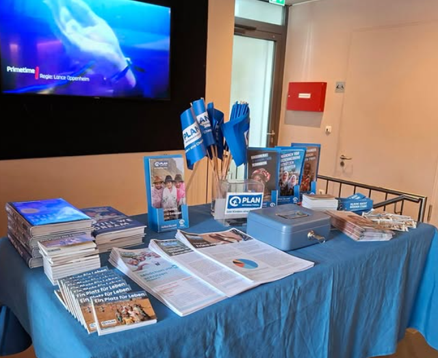 Auf einem mit einem blauen Tuch bedeckten Tisch liegen Stapel von Broschüren und Faltblättern sowie eine Spendenbox für Plan International. Hinter dem Tisch ist auf einem Fernsehbildschirm ein Filmtitel zu sehen, und auf einem Ständer sind blaue Werbefahnen gruppiert.