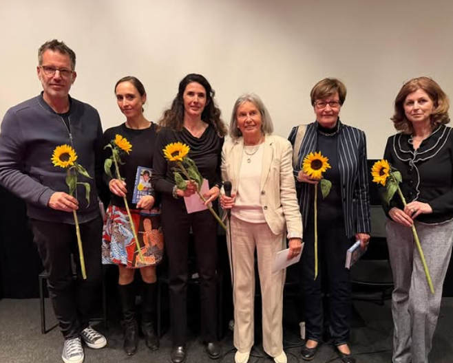 Sechs Personen stehen in einer Reihe vor einer schlichten Wand und halten jeweils eine Sonnenblume in der Hand. Sie sind leger bis elegant-leger gekleidet, wobei die zweite Person von links ein blaues Buch in der Hand hält.