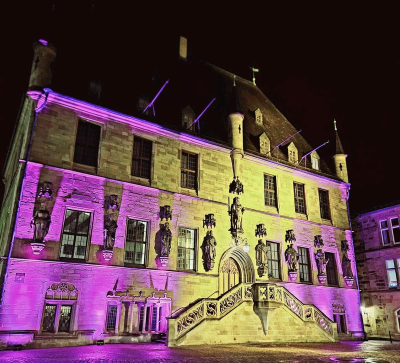 Ein altes Steingebäude wird nachts von leuchtend violetten Scheinwerfern beleuchtet. Das imposante Bauwerk besticht durch Statuen im gotischen Stil entlang der Fassade und eine kunstvoll verzierte Steintreppe, die zu einem zentralen Bogeneingang führt.