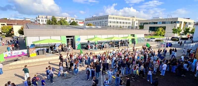 Eine große Schar von Schülern versammelt sich auf einem gepflasterten Schulplatz vor einem modernen, einstöckigen Gebäude. Grüne Sonnenschirme spenden im Eingangsbereich Schatten, und die Szene spielt sich unter einem strahlend blauen Himmel mit leichten Wolken ab.