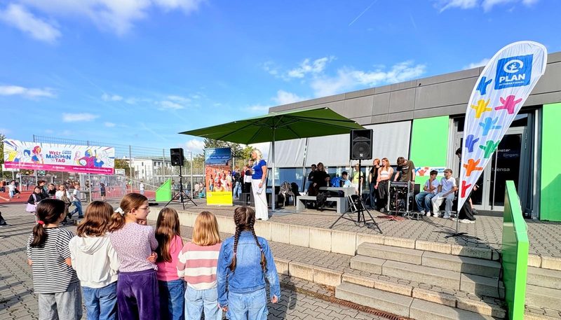 Eine Gruppe von Kindern beobachtet eine Frau, die bei einer Veranstaltung von Plan International von einer Bühne im Freien aus spricht. Die Bühne ist mit Lautsprechern, einem grünen Sonnenschirm und bunten Bannern ausgestattet und steht unter blauem Himmel, im Hintergrund ist ein modernes Gebäude zu sehen.